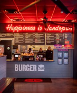 interior of Burger 25 with a neon "Happiness is Handmade" sign, a smiling staff behind the counter, and the menu displayed above.