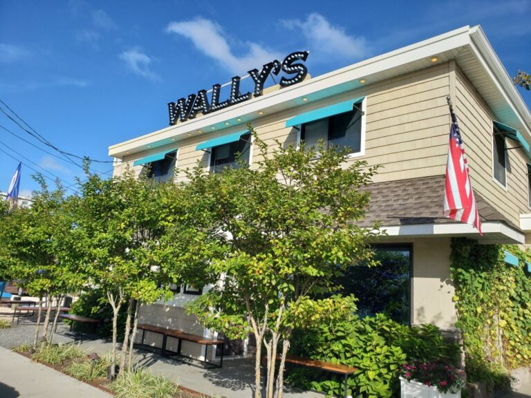 the exterior of Wally's, a brightly lit restaurant with a marquee-style sign. The building features beige siding, a row of bushes and trees in front, and an American flag hanging to the right.