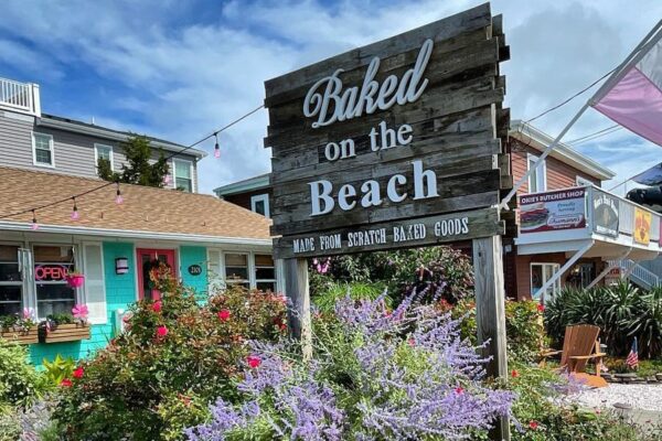 A colorful bakery called "Baked on the Beach" with a wooden sign and an "OPEN" flag outside, surrounded by flowers and coastal-style buildings