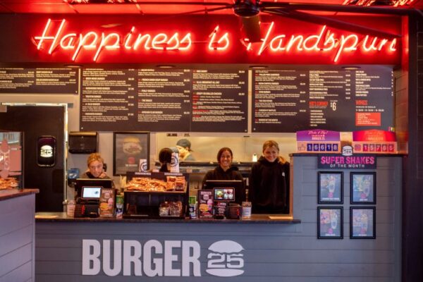 Burger 25 interior of Burger 25 with a neon "Happiness is Handmade" sign, a smiling staff behind the counter, and the menu displayed above.