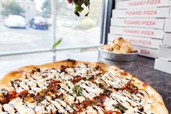 The image shows a close-up of a pizza topped with fresh ingredients, as someone is adding a sprinkle of basil on top.